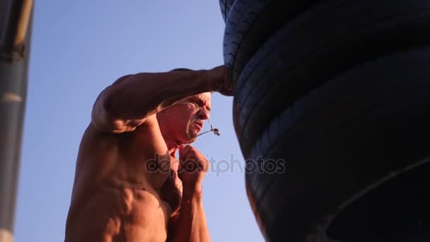 Joven atlético con torso desnudo, desnudo, boxeo, practica la técnica de las huelgas, lucha con saco de boxeo. 4k cámara lenta. en la playa, en verano, en los rayos del sol . — Vídeo de stock