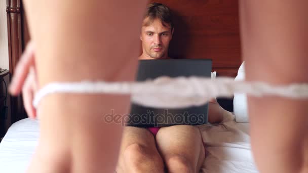 Сексуальная женщина пытается соблазнить мужчину, работающего над ноутбуком в постели. Девушка снимает нижнее белье перед своим парнем, а он игнорирует ее, работает на ноутбуке. 4k, slow motion — стоковое видео