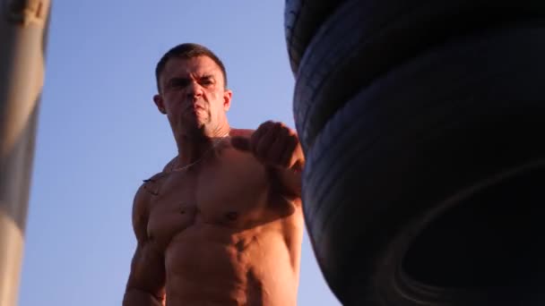 Jeune homme athlétique avec torse nu, boxe, pratique la technique des frappes, se battre avec un sac de boxe. 4k Ralenti. sur la plage, en été, sous les rayons du soleil . — Video