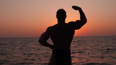 bir erkek vücut geliştirmeci silüeti. gün batımında sahilde poz güzel atlet. 4 k. yavaş çekim