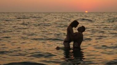 Bikini ve romantik kaslı adam hamile kadın siluetleri. vücut geliştirmeci hugs, karısını öpüyor ve karnı vuruş. Deniz günbatımı arka planı. 4 k. ağır çekim