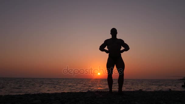 男性のボディービルダーのシルエット。夕暮れ時のビーチでポーズ美しい選手。4 k. 低速撮影 — ストック動画