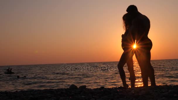 Silhouette di donne incinte in bikini e romantico uomo muscoloso. il bodybuilder abbraccia, bacia sua moglie e le accarezza la pancia. sullo sfondo di un tramonto sul mare. 4k. Rallentatore — Video Stock