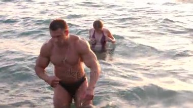 Bikini ve romantik kaslı adam hamile kadın siluetleri. vücut geliştirmeci hugs, karısını öpüyor ve karnı vuruş. Deniz günbatımı arka planı. 4 k. ağır çekim