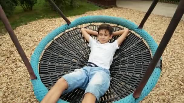 Мальчик качается на качелях в гамаке в парке развлечений в городе. 4k, пространство для копирования — стоковое видео
