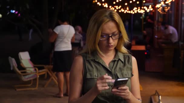 Блондинка друкує смс на смартфоні в темряві, влітку. вечір, ніч. 4k, повільний рух — стокове відео