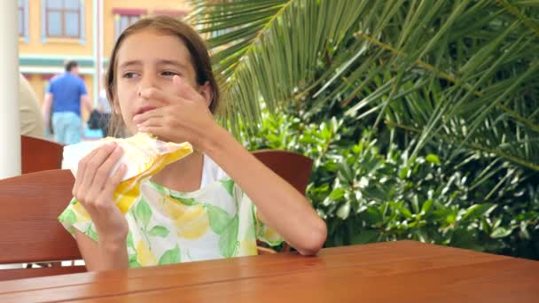 Seorang gadis yang menarik makan roti pita segar dengan salad sayuran segar dan daging, duduk di kafe makanan cepat saji di bawah pohon di taman. 4K gerak lambat — Stok Video