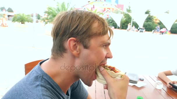 Привлекательный мужчина ест свежую хлебную питу с салатом из свежих овощей и мяса, сидя в кафе быстрого питания под деревом в парке. 4k, slow motion — стоковое видео