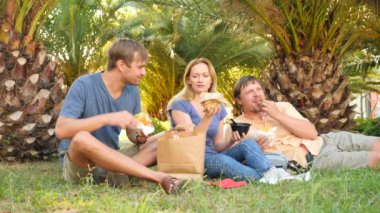 Üç arkadaş, erkek ve kadın hızlı avuç içi altında çimlerin üzerine oturup yemek yemek. 4k, ağır çekim