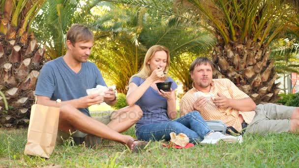 Trois amis, hommes et femmes mangent de la restauration rapide assis sur l'herbe sous les paumes. 4k, au ralenti — Video