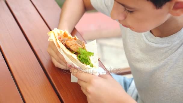 Привлекательный мальчик ест свежую хлебную питу с салатом из свежих овощей и мяса, сидя в кафе быстрого питания под деревом в парке. 4k, slow motion — стоковое видео
