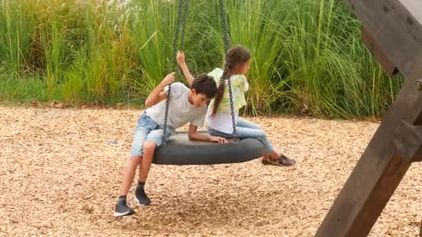 Счастливые брат и сестра, качающиеся на качелях в парке развлечений в городе. 4К, скопируйте пространство, замедленная съемка. стиль жизни — стоковое видео