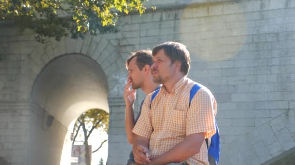 Депресивний і сумний молодий чоловік курить цигарку зі своїм другом на тлі мосту. 4k, повільна стрільба, копіювання простору. дим з цигарки на сонячному світлі — стокове відео