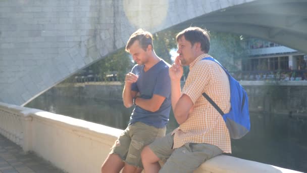 Ein deprimierter und trauriger junger Mann raucht mit seinem Freund vor dem Hintergrund der Brücke eine Zigarette. 4k, Zeitlupe, Kopierraum. Rauch einer Zigarette im Sonnenlicht — Stockvideo