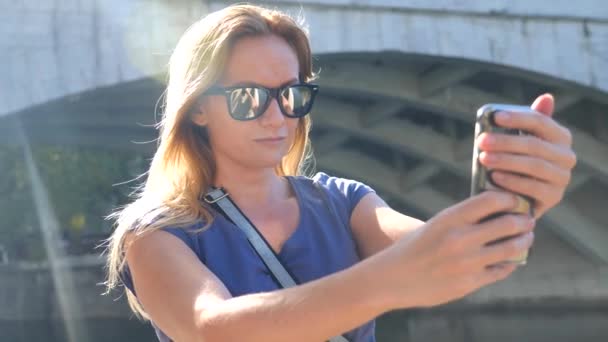 Hermosa mujer adulta joven tomando fotos de sí misma, selfie. contra el fondo del puente sobre el río. 4k, grabación en cámara lenta, espacio para copiar . — Vídeos de Stock