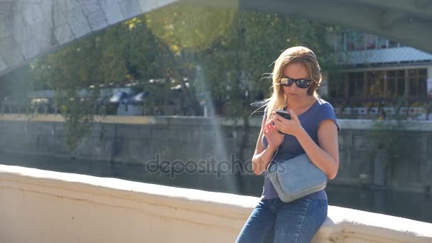 Mooie jonge volwassen vrouw nemen foto van zichzelf, selfie. tegen de achtergrond van de brug over de rivier. 4k, slow-motion-video schieten, ruimte kopiëren. — Stockvideo