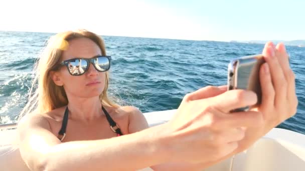 Una mujer atractiva en un yate en un día de verano en el mar se hace en su teléfono inteligente. una rubia tetona delgada con pelo largo, con gafas de sol. 4, espacio de copia, primer plano. Movimiento lento — Vídeo de stock