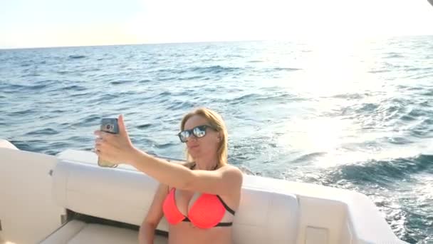 有魅力的女人，在海上的一个夏日游艇上使自己对她的智能手机。薄丰满金发女郎，有着长长的头发，戴着太阳镜。4，复制空间，特写。慢动作 — 图库视频影像
