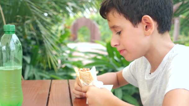 Ein attraktiver Junge isst ein frisches Fladenbrot mit einem Salat aus frischem Gemüse und Fleisch und sitzt in einem Fast-Food-Café unter einem Baum im Park. 4k, Zeitlupe — Stockvideo