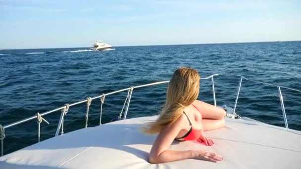 Mulher atraente em um iate em um dia de verão no mar. loira peituda magra com cabelos longos, usando óculos de sol e um biquíni vermelho brilhante está tomando banho de sol no convés de um barco em mar aberto. câmara lenta — Vídeo de Stock