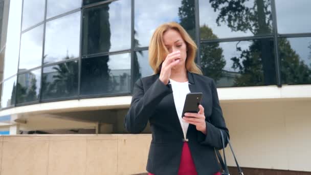 Mujer de negocios en un traje de negocios con un teléfono inteligente, caminando en un centro de negocios, caminando en la ciudad, steadicam tiro. 4k, cámara lenta, espacio para copiar — Vídeo de stock