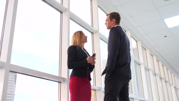 Homme et femme en costume se tiennent debout et parlent. Homme d'affaires et femme d'affaires sont dans le couloir du bureau devant la fenêtre. 4k, au ralenti — Video