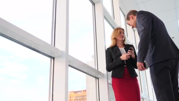 Мужчина и женщина в костюмах стоят и разговаривают. Бизнесмен и деловая женщина находятся в офисном коридоре напротив окна. 4k, slow motion — стоковое видео