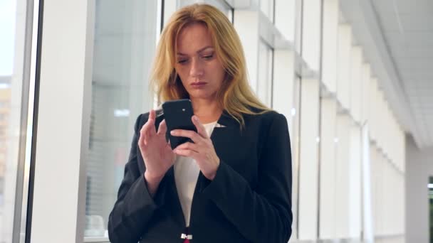 Zakenvrouw in een bedrijf passen met een smartphone, wandelen langs de corridor van het business center met panoramische ramen, 4k, langzame beweging, ruimte voor kopiëren — Stockvideo