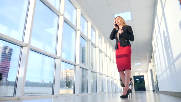 Mujer de negocios en un traje de negocios con un teléfono inteligente, caminando por el pasillo del centro de negocios con ventanas panorámicas, 4k, cámara lenta, espacio para copiar — Vídeo de stock