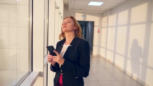 Donna d'affari in giacca e cravatta con smartphone, passeggiando lungo il corridoio del business center con finestre panoramiche, 4k, rallentatore, spazio per copiare — Video Stock