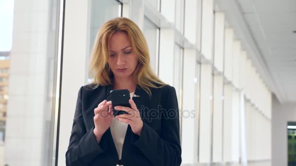 Mujer de negocios en un traje de negocios con un teléfono inteligente, caminando por el pasillo del centro de negocios con ventanas panorámicas, 4k, cámara lenta, espacio para copiar — Vídeos de Stock