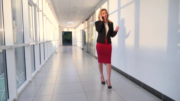 Γυναίκα των επιχειρήσεων σε μια επιχείρηση που ταιριάζουν με ένα smartphone, περπατώντας κατά μήκος του διαδρόμου από το επιχειρηματικό κέντρο με πανοραμικά παράθυρα, 4k, αργή κίνηση, χώρο για την αντιγραφή — Αρχείο Βίντεο
