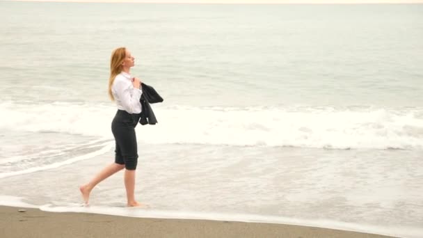 Mulher de negócios de terno em pé na praia. Ela gosta da vista para o mar. 4K, câmara lenta. ela desapertou sua camisa e inala o ar do mar — Vídeo de Stock
