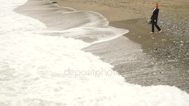 Podnikatelka v obleku, stojící na pláži. Ona má výhled na moře. 4k, zpomalené. Ona sundá jí vysoké podpatky a jde do vody — Stock video