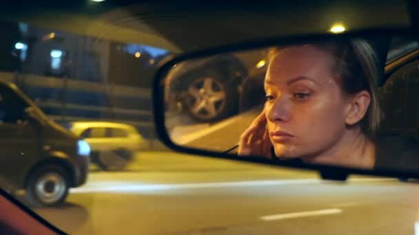Riflessione di un volto femminile con gli occhi azzurri nello specchio retrovisore, donna al volante di notte, luci notturne della città offuscate e fari dell'auto. 4k, rallentatore — Video Stock