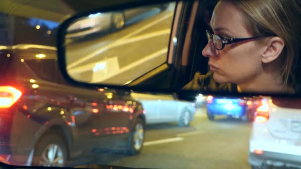 Reflexão de um rosto de mulher com olhos azuis no espelho retrovisor, mulher ao volante à noite, luzes noturnas turvas da cidade e faróis do carro. 4k, câmera lenta — Vídeo de Stock