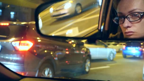 Reflejo de una cara de mujer con ojos azules en el espejo retrovisor, mujer detrás del volante por la noche, luces de noche borrosas de la ciudad y faros del coche. 4k, cámara lenta — Vídeo de stock