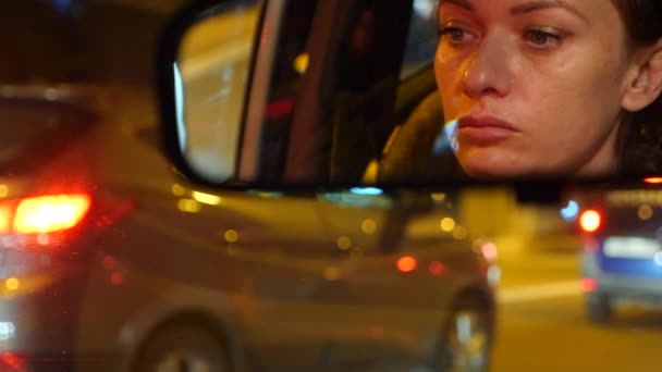 Отражение женского лица с голубыми глазами в зеркале заднего вида, женщина за рулем ночью, размытые городские ночники и автомобильные фары. 4k, slow motion — стоковое видео