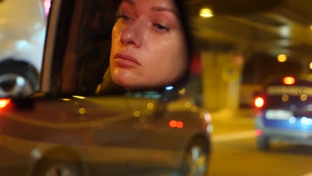 Reflet d'un visage de femme aux yeux bleus dans le rétroviseur, d'une femme au volant la nuit, de lumières de ville floues et de phares de voiture. 4k, au ralenti — Video