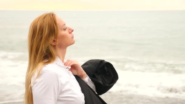 Interesu w garniturze stojącego na plaży. ona cieszy się widokiem na morze. 4k, zwolnionym tempie. ona jej koszulkę rozluźniony i wdycha powietrze morskie — Wideo stockowe