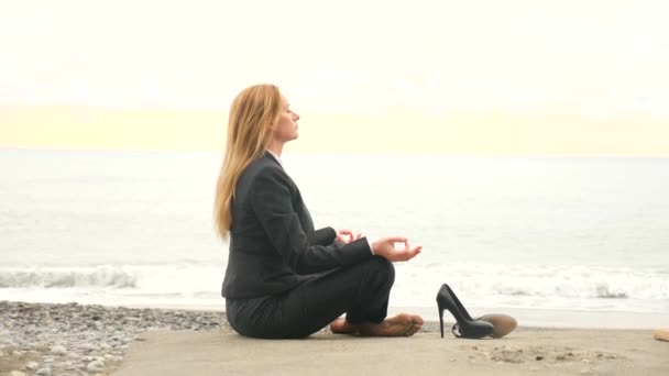 Femme d'affaires en costume sur la plage. assis en position lotus et méditant au bord de la mer. 4k, au ralenti — Video