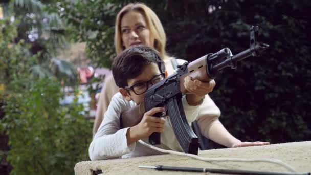 Young man shoots in a play dash from a Kalashnikov assault rifle, 4k, airsoft. family, entertainment in an amusement park. mother and her son are shooting at a dash. — Stock Video