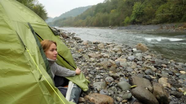 4k. 在山岸边的旅游帐篷。女孩从帐篷里爬出来, 看着那条山河. — 图库视频影像