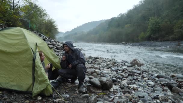 4 k. uitzicht vanaf een toeristische tent aan een rivier berg. de man met het meisje giet thee in de thermos fles en kijkt naar de rivier van de berg. — Stockvideo