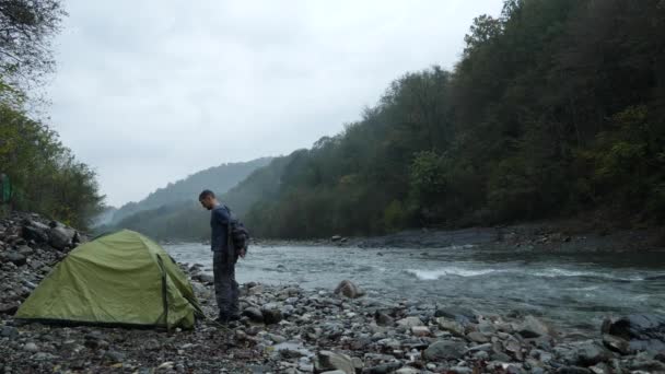 4k. un tipo cerca de una tienda de campaña turística en la orilla de un río de montaña . — Vídeos de Stock