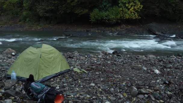 4k. el tipo pone una tienda de campaña turística en la orilla de un río de montaña . — Vídeo de stock
