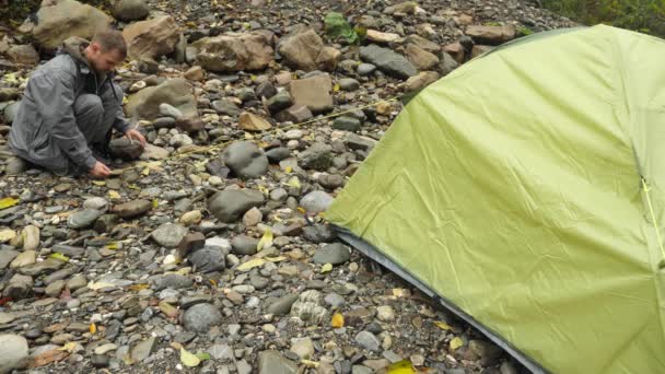 4 k. de man zet een toeristische tent op de oever van een rivier berg. — Stockvideo