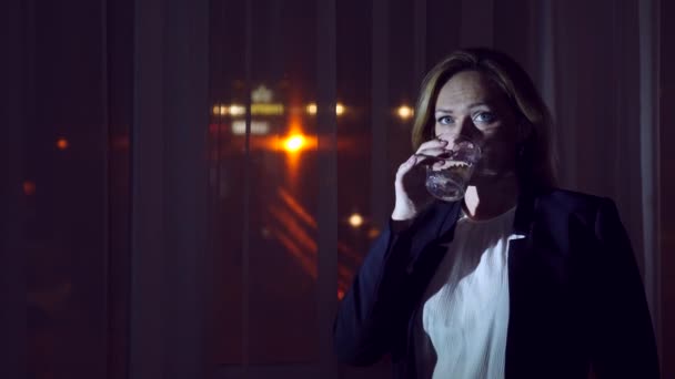 自宅窓際夜立っている飲料を飲んでビジネス スーツの若い女性。[パノラマ] ウィンドウ、街、夜景の眺め。4 k — ストック動画