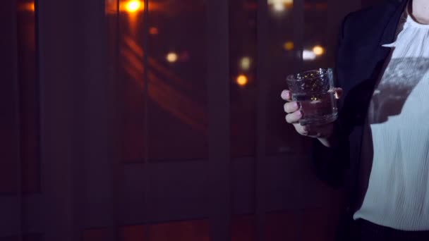 Mujer joven en un traje de negocios bebiendo bebida de pie junto a la ventana en casa por la noche. Ventana panorámica, vista de la ciudad, luces nocturnas. 4k — Vídeo de stock