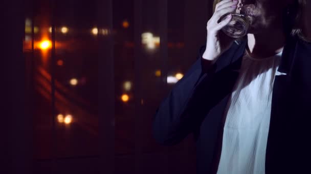 Mujer joven en un traje de negocios bebiendo bebida de pie junto a la ventana en casa por la noche. Ventana panorámica, vista de la ciudad, luces nocturnas. 4k — Vídeos de Stock